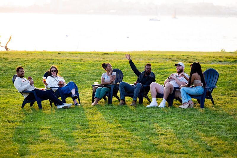The image shows a group of friends relaxing outdoors on a sunny day. They are sitting in chairs on a grassy field, with a body of water in the background. Some of them are raising their hands, and they seem to be enjoying each other's company. The atmosphere appears relaxed and cheerful.